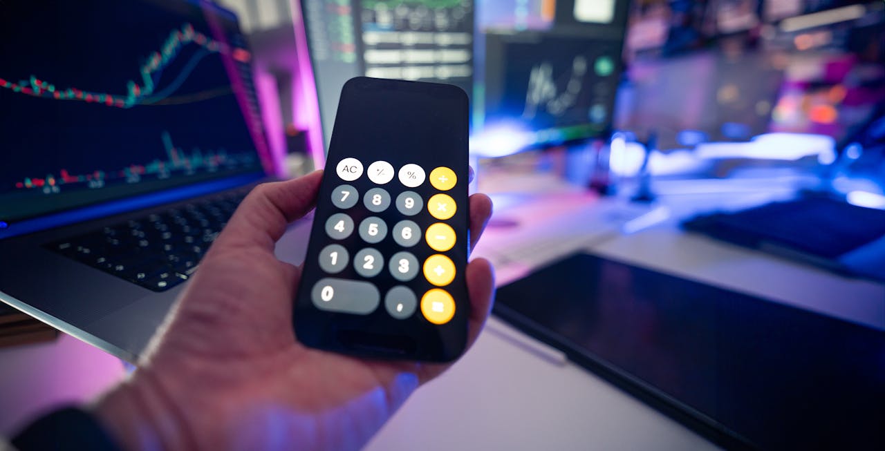 Smartphone with calculator app against financial charts on monitors, symbolizing financial planning.